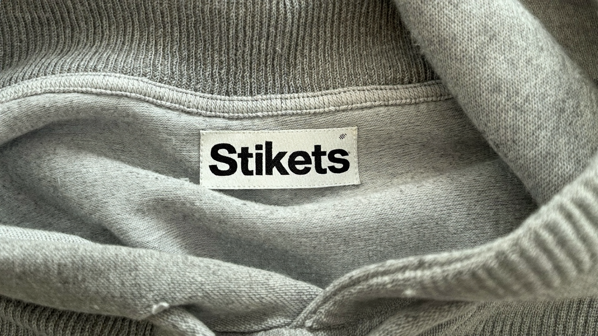 Etiqueta blanca con el nombre Stikets cosida en el cuello interior de una sudadera gris