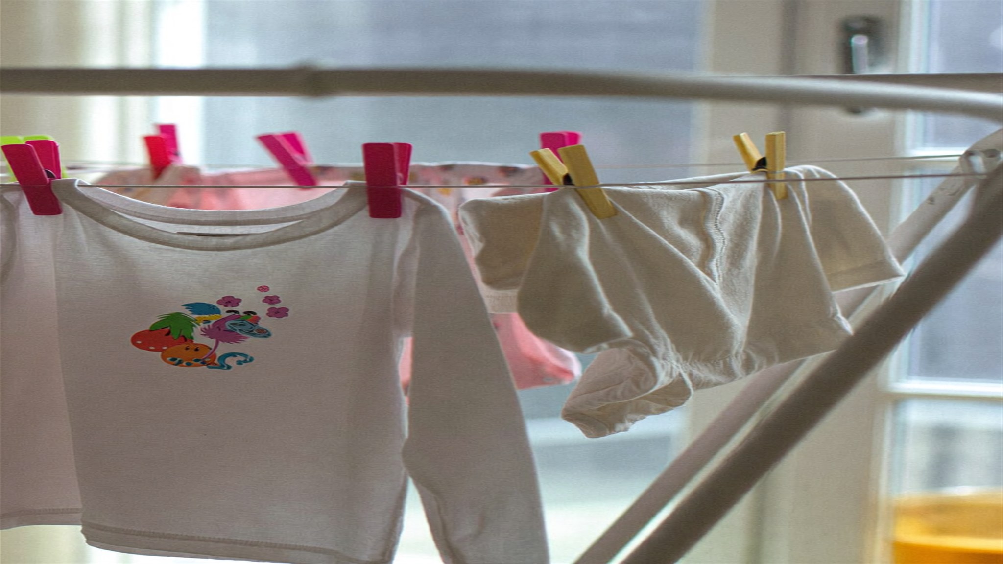 Ropa infantil blanca y de colores pastel tendida con pinzas junto a una ventana soleada