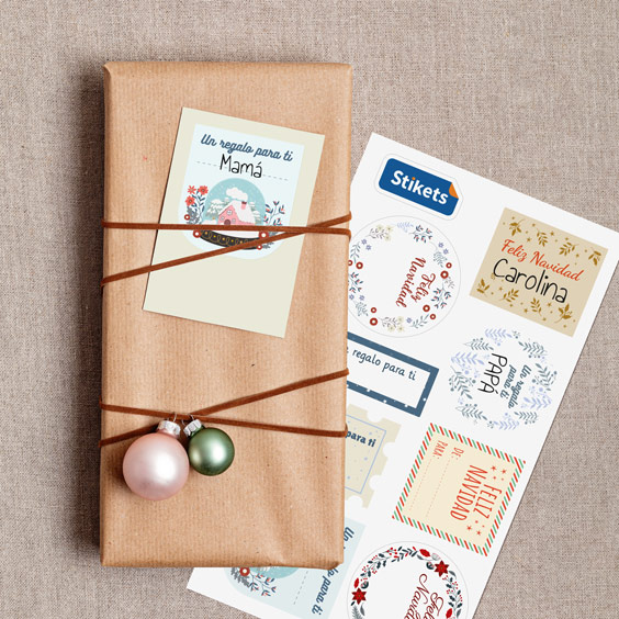Regalo envuelto en papel kraft con etiqueta navideña para escribir de Stikets junto a una lámina de diseños de Navidad